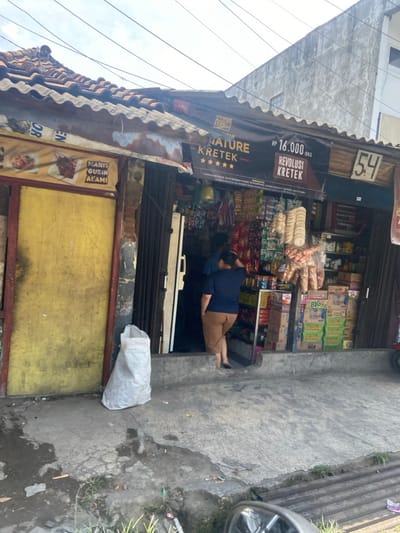 The image shows the front of a small shop in Kecamatan Dayeuhkolot, Indonesia, at latitude -6.966 and longitude 107.592. The shop is located on a street corner and has a yellow wall with a sign that reads "Manis Gurih Alami." The shop is open and a woman in a blue shirt and brown pants is walking out of the shop. There are many products on the shelves inside the shop, including snacks, drinks, and other household goods. There is a white bag on the ground outside the shop. The shop is in a residential area with a cloudy sky above.