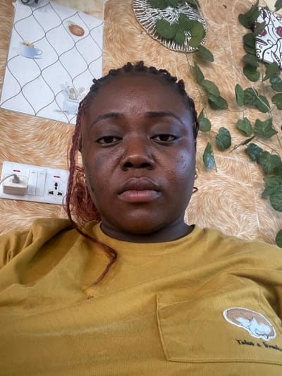 The image presents a close-up, chest-up view of an adult female centered in the frame, looking directly at the camera with a neutral expression. She has dark skin, braided hair with visible reddish-brown strands, and a small stud piercing on her left nostril. She is wearing a mustard yellow long-sleeved top that features a patch pocket on the left side of the chest, embroidered with a sleeping animal (possibly a dog or cat) and the text "Take a Break."

The background consists of a wall covered in a light brown, textured material resembling fur or a fluffy fabric. Various decorative elements adorn the wall: In the upper left quadrant, a white rectangular panel with a pattern of intersecting diagonal lines and illustrations of teacups and a single seed/bean is visible. In the upper right, a circular metallic mesh decoration is partially visible, interwoven with artificial green leaves. Below this, a long strand of artificial green ivy-like leaves hangs vertically down the wall. In the lower left quadrant, a white electrical outlet plate is mounted on the wall, featuring multiple switches and three-pin sockets, one of which has a white plug inserted. Red indicator lights are visible on some of the switches.

The specific location within the city of Abuja, Federal Capital Territory, Nigeria, cannot be determined from the visual information provided in the image.