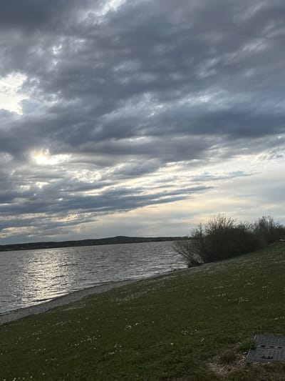 The image depicts a landscape in Gunzenhausen, Germany. On the left side of the image, a cloudy sky dominates, with the sun partially obscured by the clouds. A body of water reflects the sunlight. A grassy area is visible on the right. A sewer gate is in the grassy area.