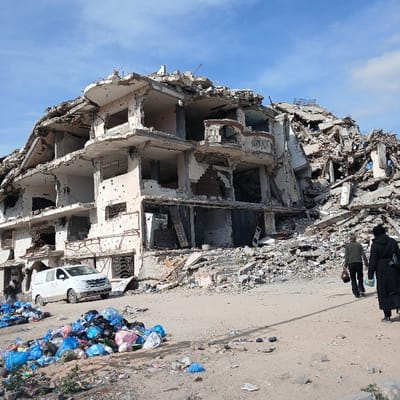 The image depicts a scene of severe urban destruction in Jabalia, Palestinian Territory, under a partially cloudy blue sky. A multi-story concrete building, heavily damaged and partially collapsed, stands in the left-center of the frame. Its upper levels are largely destroyed, revealing fractured concrete and exposed rebar. The exterior walls display numerous small circular perforations consistent with bullet impacts. To the right and behind this structure, a substantial pile of building rubble extends. On the unpaved ground in the left foreground, multiple large accumulations of colorful plastic trash bags are visible, along with scattered refuse. A white commercial van is parked adjacent to the base of the damaged building on the left. Two individuals are observed walking away from the camera on a dirt path: one male wearing dark clothing and carrying an object in his right hand, and one female in a long black garment and head covering.