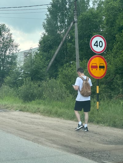 A young man wearing a white t-shirt, black shorts, white socks, and black and white sneakers stands on a dirt road. He has a brown backpack slung over his left shoulder and is looking down at his phone, which he holds in his right hand. The man is positioned to the right of two traffic signs. The first sign is circular with a red border and the number "40" in black in the center, indicating a speed limit of 40. The second sign is circular with a yellow background and a red border. It depicts two cars side-by-side with a red line through them, signifying "no overtaking." A black and yellow striped pole is visible behind these signs.
The scene is outdoors, with lush green trees and grass surrounding the dirt road. A power pole with wires stands to the left of the signs. In the background, a light-colored building structure with a metal framework is partially visible through the trees. The lighting suggests it is daytime, with a bright, overcast sky. The overall impression is a casual, everyday moment on a rural or suburban road in Novoe Devyatkino, Russia.