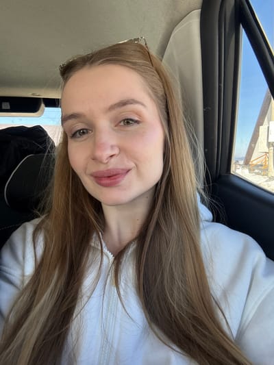 A female individual is depicted in a close-up, chest-up view, seated inside a vehicle in Virak, Montenegro. She has long, light brown hair, green eyes, and fair skin, with light pink lipstick. A pair of sunglasses with a silver-colored frame rests on top of her head. She is wearing a white zippered hooded sweatshirt. The subject looks directly towards the camera with a slight, closed-mouth smile. To her right, through the vehicle's window, a clear blue sky and a snow-covered landscape are visible, featuring a portion of an angular, light-colored structure. The vehicle's dark interior trim and light-colored headliner are partially discernible.