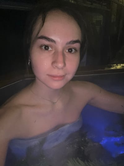 An image displays a female individual in a medium shot, partially submerged in water, consistent with a hot tub or bath setting. The environment is dimly lit, indicating nighttime or an indoor location. The water is illuminated from beneath with a blue light, which casts a blue hue on the water and reflects on the subject's skin.
The individual has fair skin, dark, short hair, and dark eyes. Water droplets are visible on her forehead, cheeks, and shoulders. She wears a small, silver-colored stud earring in her left ear and a thin, silver-colored necklace with a small circular pendant. Her expression is neutral, with a slight, closed-mouth smile, and her gaze is directed towards the camera. Her shoulders, upper chest, and right arm are visible above the water line, with the right arm extending partially into the water.
The water surface around the individual exhibits minor ripples. A dark-colored rim or edge of the water container is visible in the foreground. The background is largely dark and indistinct, offering minimal structural detail beyond the immediate water feature. No visual information within the image confirms the specified location of Базарово, Russia.