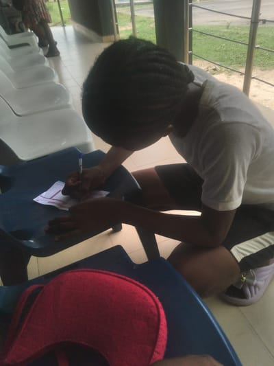 A person with braided hair and wearing a white shirt and black shorts is writing on a piece of paper resting on a blue plastic chair. The scene appears to be an indoor waiting area with rows of white plastic chairs in the background and a window showing greenery outside. The location is Osu, Nigeria.