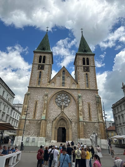 The image depicts a wide-angle, low-angle shot of the Sacred Heart Cathedral in Sarajevo, Bosnia and Herzegovina, on a cloudy day. The cathedral, a prominent Neo-Gothic structure with twin spires and stone masonry, dominates the frame. Several people are visible in the foreground and mid-ground, appearing to be tourists or locals gathered in the square in front of the cathedral. Some individuals are standing, some are seated at outdoor cafe tables, and others are walking. Architectural elements of surrounding buildings are visible on either side, including a building with white facade and neoclassical detailing to the left, and another to the right with similar detailing and awnings of outdoor seating areas. The sky is a mix of blue and white clouds.