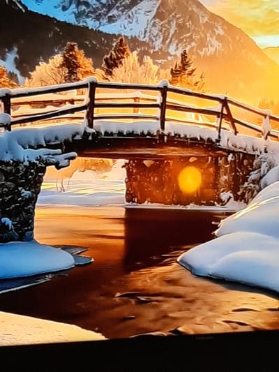 The image depicts a digitally rendered winter scene with a bridge covered in snow. The bridge is supported by stone pillars and has a wooden railing also dusted with snow. Below the bridge, a body of water reflects the warm orange and yellow light of a setting sun. The background features snow-covered trees and a large, majestic mountain under a gradient sky of blue and orange. The scene is set in the city of Sambir, Ukraine. There are no people or animals visible in the image. The overall atmosphere is serene and tranquil.
