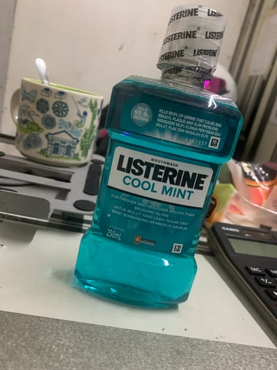 A close-up, low-angle shot captures a bottle of Listerine Cool Mint mouthwash as the primary subject, positioned on a grey textured surface. The bottle is predominantly teal in color, with clear liquid visible within, and features prominent white lettering. The cap of the bottle is wrapped with clear plastic adorned with repeating "LISTERINE" branding. To the left of the mouthwash bottle, a portion of a ceramic mug with a blue and white patterned design is visible. A silver spoon rests inside the mug. In the background, the scene is slightly blurred, showing a black calculator with a green digital display on the right side and an out-of-focus white and grey object behind the mouthwash bottle on the upper right. The overall environment suggests an indoor setting, possibly a desk or countertop. The presence of a calculator and office-like background elements implies a workspace. The image does not contain any explicit indicators of the city of Angeles, Philippines.