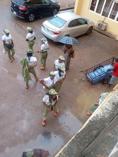 In Alagba, Nigeria, an overhead shot captures a rainy day scene in what appears to be a courtyard or open area. The ground is wet and muddy, with several large puddles reflecting the overcast sky. A black SUV is parked in the upper left of the frame, next to a silver sedan that has a license plate with "ABC-22AGH" visible.
A group of individuals, mostly young women, are scattered throughout the area, many wearing matching green khaki uniforms and white t-shirts with "NYSC" printed on them, indicating they are likely National Youth Service Corps members. Some are holding umbrellas, and others appear to be walking or standing around.
One woman in a patterned dress is holding a dark umbrella and carrying bags. A handcart filled with what appear to be bottled water or similar items is being pushed by a man in a red shirt. Another person in a red shirt is visible on the far right, looking towards the handcart. The overall atmosphere suggests a typical day, possibly with some disruption due to the weather.