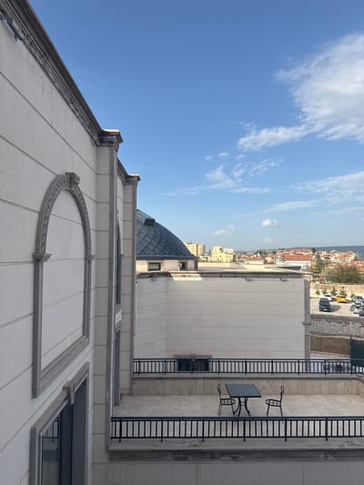 This is an exterior shot of a white stone building in Ürgüp, Turkey, with a balcony furnished with an iron table and two chairs. The building is multi-storied, featuring a domed roof and arched decorative elements. A fire escape ladder is attached to the building's side. In the background, the cityscape of Ürgüp is visible under a partially cloudy blue sky. Cars are parked on a street near the building. The photo is taken from a low angle, emphasizing the building's height.