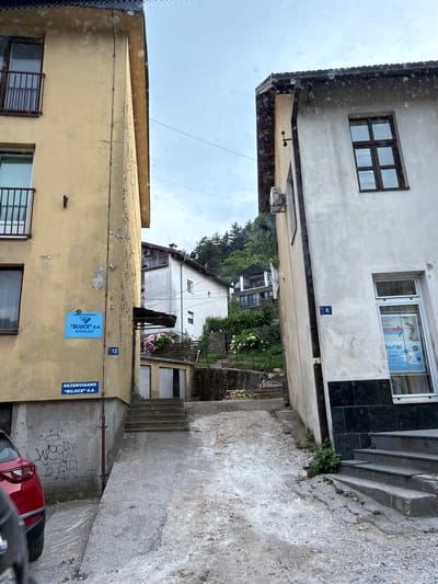 This media file is a photograph taken from a low vantage point, looking up at a narrow alleyway between buildings. The scene appears to be in Konjic, Bosnia and Herzegovina, suggested by text on signs.
The dominant subjects are the architectural elements of the buildings. On the left, a pale yellow building with peeling paint occupies the foreground. It has barred windows and some detached pieces of plaster on its facade. A blue sign on this building reads "REZREVISANO 'BUJICE' d.d.". Above this, another blue sign with white text reads "BUJICE" d.d. KONJIC". A house number, "12", is also visible.
To the right of the alley, a lighter-colored building rises higher. It features a dark tiled section near the top, a window displaying advertisements, and a smaller window above it. The building has a dark, flat roofline. A set of concrete stairs ascends along the right side of the alley, with some greenery growing in cracks.
In the background, through the alley, more buildings are visible, including a white house with a pitched roof and multiple windows, nestled amongst trees and foliage. The sky is overcast, suggesting cloudy weather. The overall impression is one of a somewhat aged urban environment, possibly a residential or commercial area. There are no people visible in the frame. The photograph seems to have been taken during daylight hours.