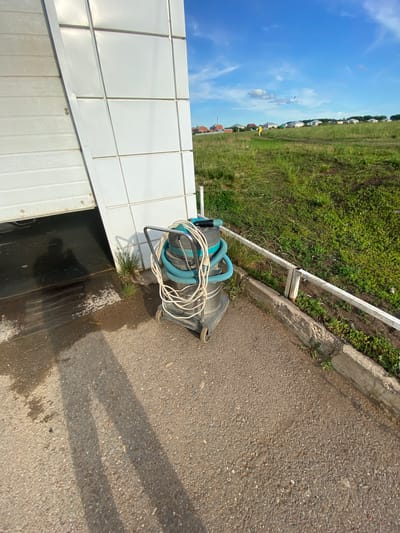 The image depicts an outdoor scene in the city of Dyurtyuli, Russia, likely on a sunny day with a clear blue sky and scattered white clouds. In the foreground, a grey industrial vacuum cleaner with blue hoses and white power cords wrapped around it rests on a paved surface. The vacuum cleaner is on wheels and has a metal handle. Adjacent to the vacuum cleaner is a large, white, sectional garage door, partially open, revealing a dark, wet interior. The ground in front of the garage door is wet and appears to be concrete with some patches of dirt and grass. To the right of the vacuum cleaner, a low white metal fence runs along a concrete curb separating the paved area from a grassy field. The field is lush and green, with some dirt paths winding through it. In the middle ground, in the distance, a person wearing a bright yellow jacket is visible walking across the grassy field. Further in the background, a row of houses with red and grey roofs lines the horizon, suggesting a residential area or the outskirts of a town. The overall environment suggests a utilitarian or industrial setting adjacent to a more natural, open space.