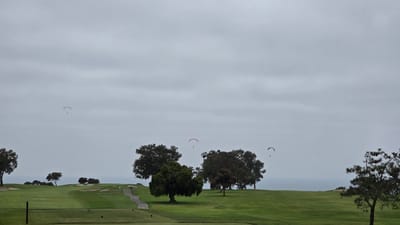 This image captures a serene view of a golf course with paragliders in the sky. The location is Torrey Pines Golf Course, in San Diego, California, USA, known for its scenic coastal setting. The sky is overcast with a thick layer of clouds, creating a diffused light. Four paragliders, each with a distinct color, are visible in the distance, suspended in the air. One paraglider is a light green, another is red, and two are blue. They appear to be enjoying a leisurely flight over the golf course.
The golf course itself is lush and green, with rolling fairways and sand traps strategically placed. A paved path winds its way through the course, suggesting a walking trail for golfers. Several trees are scattered across the landscape, adding to the natural beauty of the scene. In the background, the ocean can be seen, its surface calm under the cloudy sky, hinting at the proximity of the coastline. The overall atmosphere is tranquil and picturesque, a blend of recreational activity and natural splendor.