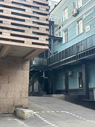 The image depicts an outdoor architectural scene in Zhabkino, Russia. The foreground features a rough-textured concrete block and a ramped asphalt surface with white dashed line markings, suggestive of a parking or loading area. To the left, a large structure with a facade of horizontal wooden slats and some exposed concrete forms the immediate environment. Extending from this structure is an overhang with wooden beams.
Dominating the right and central portion of the image is a multi-story building with a distinctive light blue brick facade. This building features multiple windows with white frames and visible air conditioning units. A metal walkway with vertical railings runs along the building's exterior at different levels. A dark metal staircase and structural supports are visible between the wooden-clad building and the blue brick building. The overall impression is an urban or industrial setting with a mix of modern and potentially older architectural elements. No people are visible in the scene.