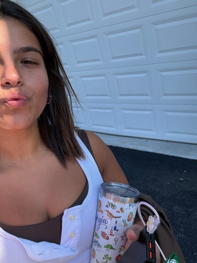 The image depicts a close-up, eye-level perspective of a female individual captured outdoors in bright daylight. The subject, positioned to the left of the frame, has dark hair and light brown skin. Her lips are pursed in a "kissy face" expression, and she wears a small hoop earring in her visible left ear. She is dressed in a white ribbed tank top with a button placket, worn over a dark brown undergarment.
In her right hand, she holds two primary objects: a white insulated travel tumbler with a clear lid and a dark rectangular object, possibly a smartphone or power bank. The tumbler is decorated with colorful illustrations, including birds, a lighthouse, a golf flagstick, a football, and palm trees. The word "Georgia" is prominently displayed in blue cursive text on the tumbler. A white coiled cable with a small, clear or white floral-shaped charm is attached to the rectangular device.
The background consists of a white paneled garage door, which occupies the upper right portion of the image. Below the garage door, a concrete strip and dark asphalt pavement are visible. The exact location within the city of Princeton, United States, is not discernible from the visual information provided in the image.