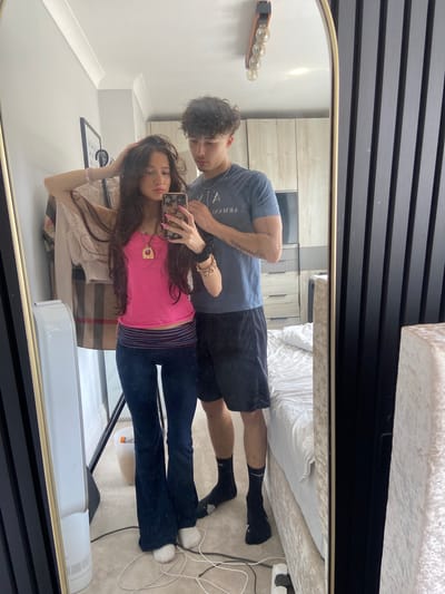A young woman and man take a mirror selfie in a bedroom. The woman is on the left, facing the mirror, with her left hand on her head. She is wearing a bright pink tank top, dark flared pants, and white socks. She has long, dark brown hair and is wearing a gold necklace. The man is behind and to the right of the woman, also facing the mirror. He is wearing a blue t-shirt, black shorts, and black socks. His left arm is around the woman's waist. The room appears to be a bedroom with a bed covered in white linens, a wardrobe, and a dresser visible in the mirror's reflection. A fan is also visible to the left of the couple. The overall impression is of a casual, domestic setting. The location is believed to be Welwyn Hatfield, United Kingdom.