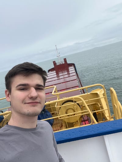 A single young adult male with dark hair and light complexion is depicted in a selfie, looking directly at the camera with a faint smile. He is wearing a grey long-sleeved shirt. The subject is positioned on the deck of a vessel, with a blue railing, wet with water droplets, in the lower right foreground. Behind and below the subject, the aft section of a cargo ship is visible, featuring multiple red, reflective cargo hatch covers and yellow metallic structures associated with the ship's equipment. The vessel is situated on open water, under an overcast, grey sky. A low-lying landmass is discernible on the distant horizon to the right. The scene is consistent with a maritime location near Bangor, United Kingdom.