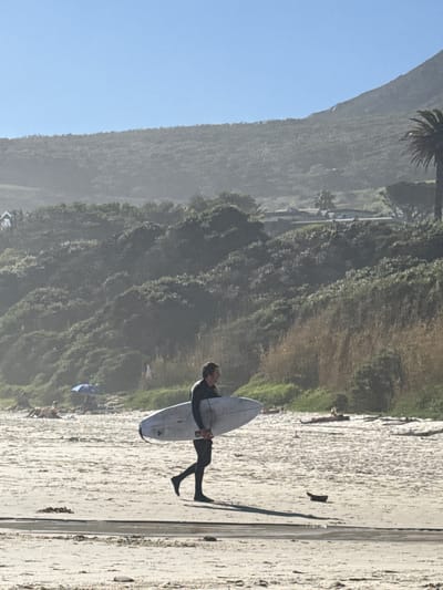 A surfer in a wetsuit holds a surfboard on a sandy beach in Hout Bay, South Africa. A mountain covered in green vegetation rises in the background under a clear blue sky. A small group of people are visible in the distance.