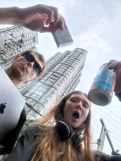 A low-angle, upward-facing urban snapshot captures two individuals in what appears to be the Jing'an District, China, under an overcast sky. In the foreground, a male subject with black sunglasses is smoking a cigarette, holding a silver laptop (likely a MacBook, indicated by the Apple logo) with his right hand. Above his left hand, he holds a red and white packet of cigarettes, on which Chinese health warnings are clearly visible: "尽早戒烟有益健康" (Quit smoking early for health) and "戒烟可减少 对健康的危害" (Quitting smoking can reduce harm to health).
To the top-right, a female subject with long, light brown hair wears black over-ear headphones around her neck and displays a surprised or expressive open-mouthed facial expression. She holds an upside-down blue Monster Energy drink can, with its distinctive logo and text visible, above her head.
The background is dominated by modern, tall high-rise buildings with many windows, characteristic of an urban cityscape. Overhead utility lines and poles are also visible, further establishing the metropolitan setting. The image uses an unconventional perspective, focusing on the subjects and their personal items against the backdrop of the city on a cloudy day.