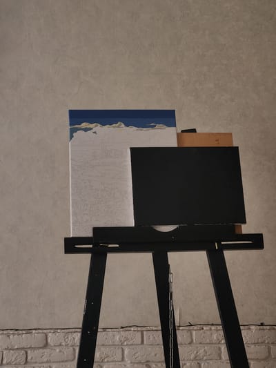 An art easel stands in front of a textured wall. On the easel, a canvas displays a paint-by-numbers image of a sky with clouds and a landscape with numbered outlines. To the right of the canvas is a black board. The easel is made of black wood and has a chain hanging from it. In the background, a white brick wall is visible at the bottom. The scene is indoors, and there are no people present. The location is not specified as Murom, Russia; no identifiable landmarks or context suggest this location.
