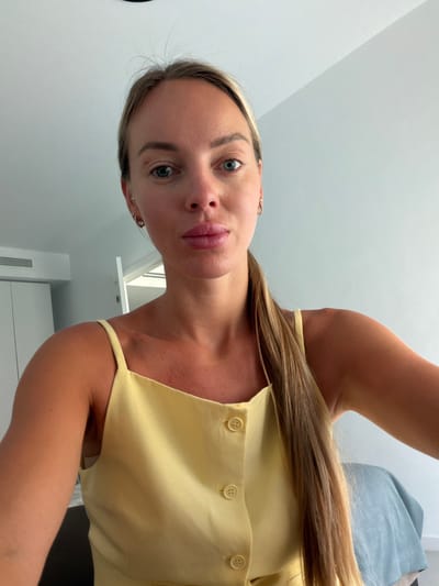 A fair-skinned Caucasian woman with blonde hair pulled back in a ponytail and blue eyes is looking directly at the camera. She is wearing a yellow tank top with buttons down the front. The background appears to be a room with white walls and a white ceiling. There is a closet and an air conditioning unit visible on the left side of the image. The lighting is bright and natural, suggesting it is daytime. The scene appears to be an indoor setting in an apartment or hotel room in Finestrat, Spain.