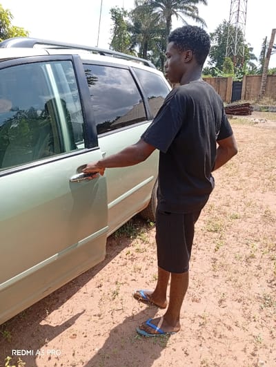 The image depicts an outdoor scene in Osina, Nigeria. A single male subject with a dark complexion is standing in profile, facing the right, next to the passenger side of a light-colored (possibly light green or beige) minivan or SUV equipped with roof rails. He is wearing a black short-sleeved t-shirt, black shorts, and blue and light blue flip-flops. His right hand is grasping the chrome handle of the vehicle's rear passenger door, suggesting an action related to opening or closing the door. The ground is unpaved, consisting of dry, reddish-brown earth with sparse green vegetation. Shadows are cast on the ground, indicating direct sunlight. In the background, several palm trees and other leafy trees are visible. To the right, a metal lattice communication tower stands prominently. A wall, possibly concrete or brick, and a dark gate structure are also present in the background.