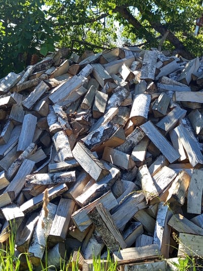 This media file depicts a large stack of cut firewood, primarily birch, piled outdoors in Ostrovtsy, Russia. The logs are of various sizes and lengths, with some showing the distinctive white bark of birch trees. They are arranged in a haphazard yet sturdy manner, indicating they have been stacked for storage. The scene is illuminated by natural sunlight, suggesting it is daytime, likely during a sunny period given the strong shadows cast by the wood.
The background features the lush green foliage of trees, with sunlight filtering through the leaves, creating dappled light effects on the firewood. To the right side of the image, blades of green grass are visible, further confirming an outdoor setting. The overall impression is one of preparation for cooler weather, with a substantial amount of wood gathered and stacked. There are no people visible in the image, and the focus is entirely on the objects and the natural environment. The visual cues, such as the type of wood and the surrounding vegetation, are consistent with a rural or semi-rural location. No text is visible in the media file.