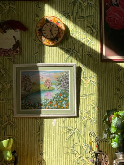 The image displays a wall decorated with bamboo-patterned wallpaper. Several framed items are hung on the wall. In the upper left corner, a framed artwork of a red rose is partially visible. To its right, a cluster of green plants hangs, with a decorative jewelry stand holding various trinkets. Below the rose artwork is a round clock with Roman numerals and a festive wreath design. Center and slightly to the right is a framed embroidery of a landscape scene featuring a body of water, trees, and flowers, with a faint silhouette of a bird. Below the clock, a framed piece with decorative elements and some handwritten text is visible. A portion of a green desk lamp is in the bottom right corner. Sunlight casts diagonal shadows across the wall, suggesting it is daytime. The overall impression is of a home interior, possibly in a residential setting in Гаспринский поселковый совет, Ukraine. No specific activity or event is depicted; the scene appears to be a static display of decorative items. There is no discernible text in the image, other than the numerals on the clock face.