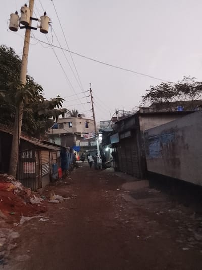 A narrow, dirt road runs through a densely populated area. Buildings on both sides appear to be a mix of residences and small shops. Overhead, a network of power lines stretches across the sky. A utility pole with transformers stands prominently on the left. The scene is dimly lit, suggesting early morning or late evening. Debris and garbage are scattered along the roadside, indicating the area may be impoverished. A few people walk in the distance.