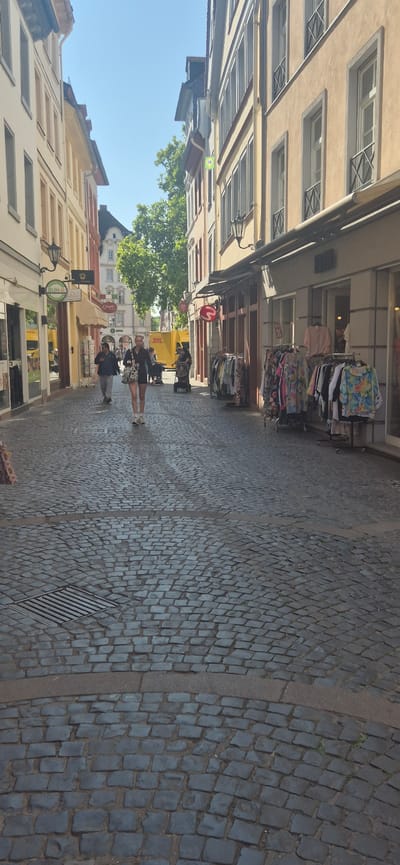 The image shows a narrow cobblestone street in Mainz, Germany, lined with traditional European buildings. A few people are walking along the street. In the foreground, the cobblestone pavement is detailed, with a metal grate on the left. Several shops with awnings are visible on both sides of the street. Clothes are displayed on racks outside some of the shops. A yellow DHL truck is parked further down the street. The sky is clear and blue, indicating a sunny day.