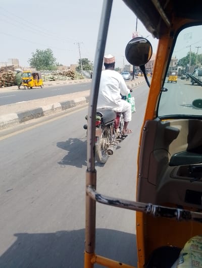 The image depicts a daytime street scene in Dorayi, Nigeria, observed from the interior of a yellow-orange vehicle, likely a tricycle. A male individual, viewed from behind, is operating a dark-colored motorcycle on a paved road, wearing a white tunic, a light-colored cap, and sandals. A green and white patterned bag hangs from his motorcycle. The road features a concrete median barrier with black and white stripes. On the left side of the road, a person in green attire stands next to a yellow tricycle, with large stacks of cut timber or logs piled behind them. In the distance, other vehicles including cars and another yellow tricycle are visible on the multi-lane road. Overhead power lines, poles, scattered trees, and buildings are present in the background under a clear, bright sky. Shadows indicate strong sunlight.