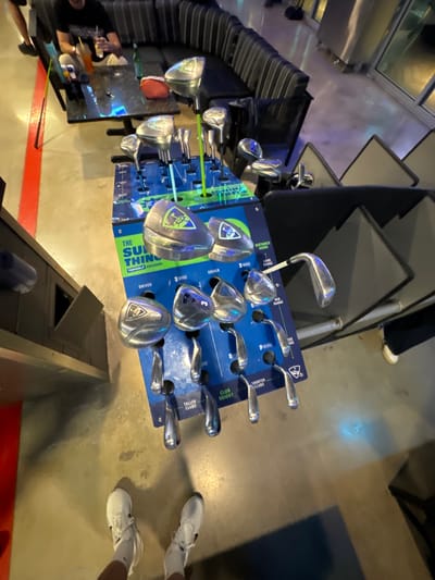 An interior photograph depicts a retail and social area at Topgolf Miami Gardens, located at 17321 NW 7th Ave, Miami Gardens, Florida, United States.
In the midground, a prominent blue and green vertical display stand showcases approximately 25 silver-colored golf clubs. The stand is labeled "THE SUM OF THINGS" and "TOPGOLF EXCLUSIVE," with sections for drivers, 3-woods, and irons (numbered 8, P, S), categorized by "CLUB HEIGHT" and "DISTANCE GUIDE" (long, mid, short range). Several club heads feature a green shield logo.
In the upper left background, one individual is seated on a black, ribbed sectional sofa. This person wears a dark short-sleeved shirt and holds an amber-colored drink with a straw in their right hand. A similar drink and a small red pouch are present on a dark, reflective rectangular table in front of the sofa.
To the far right background, the lower legs and white athletic shoes of another individual are partially visible. Beyond this, a reflective surface, likely a large window or architectural element, shows ambient light.
The floor is a light gray concrete. A red stripe delineates a boundary along the left side of the frame. In the immediate foreground, the photographer's feet, clad in white athletic shoes and white socks, are visible, indicating an overhead perspective directed towards the golf club display.