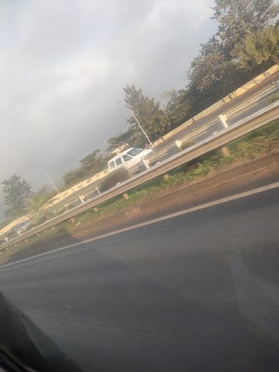 The image depicts an exterior view of a highway or major road in Theta ward, Kenya, captured from a moving vehicle's interior. The perspective is angled slightly upwards from the lower left.

In the immediate foreground, dark asphalt pavement is visible, with a solid white line marking a lane edge. White numbers "4130" are stenciled on the asphalt surface near a metallic guardrail.

A silver-colored, four-bar guardrail extends horizontally across the midground, separating the foreground lane from an elevated or adjacent section of the road. Beyond the guardrail, a light-colored concrete barrier wall features partially legible white text. Behind this barrier, a multi-lane road is visible. A white van or SUV is traveling away from the viewer on this road. Further down, a dark sedan is partially visible. A single individual, possibly wearing a red top, is situated on the far side of the road near the white van, either standing or walking.

The background consists of dense green foliage from trees and shrubs lining the roadside, punctuated by tall streetlights. The sky is uniformly overcast and light grey, indicating diffuse lighting conditions.