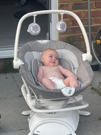 A baby is lying in a grey and white striped baby bouncer outdoors. The baby is wearing a diaper and is looking up and to the left, with a slight smile. The bouncer has a white frame and two hanging plush toys, one on each side, with images of a bear's face. The bouncer is positioned on a paved surface, possibly a patio or driveway, with some grass and brickwork visible in the background. A yellow flower is growing near the brickwork. The lighting suggests it is daytime, likely sunny. The overall scene is casual and domestic. There is no visible text.
