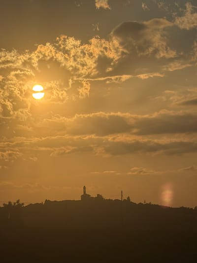 The image shows a view of Santo Stefano Belbo, Italy, at sunset or sunrise. The sun, partially obscured by clouds, is positioned in the lower left of the frame. The sky is filled with clouds, some in streaks and others in more scattered formations. The silhouette of the town's skyline is visible along the right edge of the image. A distinct tower-like structure is identifiable in the skyline. The overall lighting suggests a hazy or dusty atmosphere.