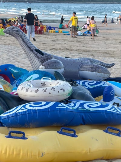 In Cam Lam, Vietnam, a pile of inflatable pool toys rests on a sandy beach, with the ocean and a few distant beachgoers visible in the background. The foreground is dominated by the colorful inflatables, including a large, gray dinosaur-shaped float, several blue and white swirl-patterned tubes, a light-colored tube with rabbit graphics, and a yellow tube with blue handles. Some of the inflatables appear worn and dusty from use. In the middle ground, people are scattered on the sand, some sitting at tables under umbrellas, while others are walking or standing near the water. The sea is calm with gentle waves.