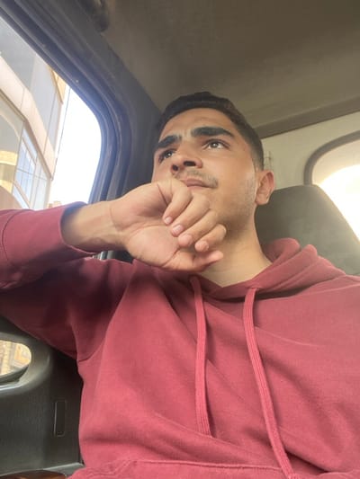 The image displays a young adult male inside a vehicle, positioned slightly off-center and looking upwards and to his left. He has short, dark hair, dark eyebrows, and a light mustache. He is wearing a dark red or maroon hooded sweatshirt with visible drawstrings. His right hand is raised, with fingers curled and knuckles near his mouth or chin. The vehicle's interior features dark plastic or fabric elements on the door frame and ceiling. Through the open window on the left side of the frame, a light-colored building with multiple rectangular windows is visible under daylight conditions. No specific identifying landmarks within the image confirm its exact location in the city of Ma'raba, Syria.