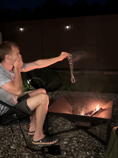 In Starokorsunskaya, Russia, a man sits in a black patio chair next to a square metal fire pit containing a small fire. He is wearing a gray t-shirt, gray shorts, and black Crocs. He is holding a long, beaded object over the fire, from which smoke is emanating. Behind him is a dark fence with two lights mounted on it. The ground is covered in small grey stones.
