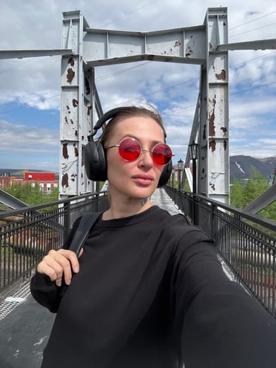 This media file is a selfie taken outdoors on a cloudy day, likely during daylight hours. The subject is a person with short hair, wearing a black long-sleeved shirt, black headphones, and distinctive round red-tinted sunglasses. They have a nose ring and visible neck tattoos. The person is holding a black bag strap.
The setting appears to be a bridge or elevated walkway constructed with weathered, peeling metal beams and a metal grate walkway. In the background, there is a view of industrial buildings, including a prominent red-roofed structure, and lush green trees, suggesting a mixed industrial and natural environment. Further in the distance, mountains can be seen, partially obscured by clouds. The sky is filled with a mix of white clouds and blue sky.
The overall mood is casual and perhaps adventurous, with the subject appearing relaxed and engaged with the camera. The location context of Norilsk, Russia, is consistent with the imagery of industrial infrastructure and mountainous terrain. No visible text is present in the image.
