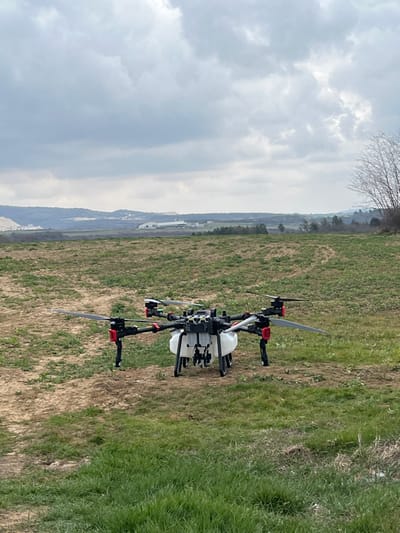 A large, multi-rotor agricultural spraying drone is positioned on an uneven field of sparse green grass and exposed soil. The drone features a black frame with red accents on its motor mounts, eight propellers (four visible), and a prominent white liquid tank beneath its central body, supported by black landing gear. In the background, a rural landscape unfolds under an overcast sky with gray and white clouds. Distant hills are visible, with a large white quarry or excavation site present on a hillside to the left. Further to the right, a long, white industrial or agricultural building is situated amidst the landscape. Bare-branched trees are partially visible on the far right of the scene. No individuals are present. The scene is located in Ruska Bela, Bulgaria.