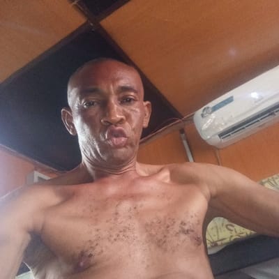 The image depicts a close-up, head-and-torso view of a shirtless adult male of apparent African descent, with a shaved head and dark complexion, making a puckered lip expression directly at the camera. Sparse dark hair is visible on his chest. The background reveals an indoor setting with an orange-brown panel ceiling and an orange-painted wall. A white wall-mounted air conditioning unit, partially wrapped in plastic or tape, is affixed to the upper right wall. Exposed electrical wiring runs along the wall and ceiling near the unit. A portion of patterned fabric, consistent with upholstery or a pillow, is visible in the lower right corner. The specific city location of Koko, Nigeria, is not discernable from any visual elements within the image.