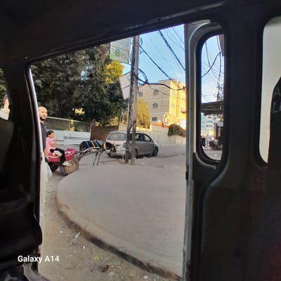 In Nuseirat, Palestinian Territories, a view from inside a vehicle captures a street scene. A young child in a pink shirt and white sneakers sits on a red seat attached to a cart pulled by a donkey. Beside the cart, a man stands, looking to his left. Behind them, a white van with Arabic writing on its rear window is parked, followed by a silver car. Overhead, power lines crisscross against a blue sky, with a light-colored building in the background. The road surface is asphalt, with some debris scattered on the ground. The image is framed by the interior of the vehicle from which it was taken, with dark surfaces along the edges.