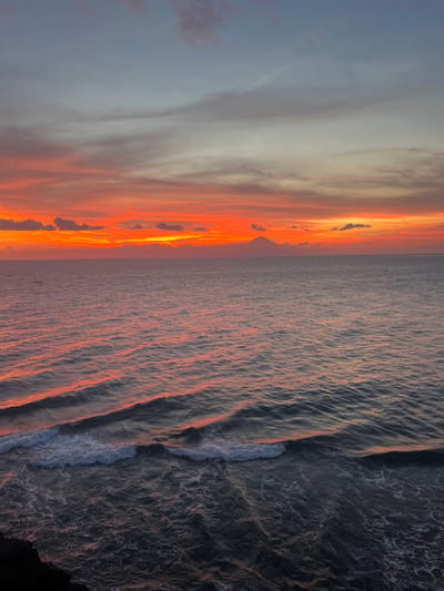 An elevated view captures a seascape at sunset in Desa Batu Layar Barat, Indonesia. The sky occupies the upper third, transitioning from a light blue-grey top to a vibrant horizon band of orange, red, and yellow, indicative of a low sun position. Scattered stratiform and cumulus clouds are present, reflecting the intense sunset colors. The horizon line is clear and flat. The ocean dominates the lower two-thirds, its surface reflecting the sunset hues in streaks of orange and pink across the water. Multiple parallel waves are visible, moving towards the foreground. The water appears dark blue-grey, intensifying in color towards the bottom. White foam caps are present on breaking waves in the immediate foreground. On the distant horizon, a dark, distinct, triangular silhouette of a mountain or volcano is visible, positioned slightly right of center. The extreme bottom left corner shows an indistinct dark mass, consistent with a cliff edge or elevated ground from which the image was captured. No individuals, artificial structures, or overt actions are present.