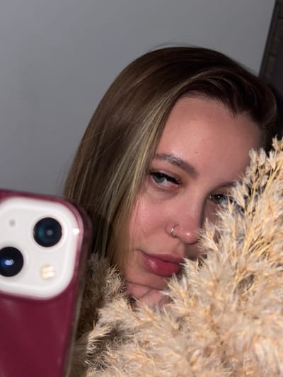 A young, light-skinned woman with blonde-streaked light brown hair is visible from the left side of her face to the center, and from her forehead to her lips. She has light grey-green eyes, a nose ring, and pinkish lips. She is holding a bundle of dried grasses or reeds, which are partially obscuring her face. The woman is holding a maroon-colored smartphone with a dual-lens camera in front of her. The background is a muted grey wall. The location is not identifiable as Port-Katon, Russia based on the image alone.