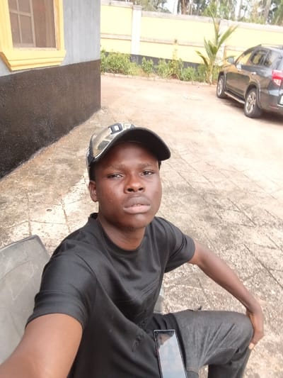A young male is depicted seated outdoors, captured in a selfie-style photograph. He has a dark complexion and wears a black short-sleeved t-shirt, dark grey athletic pants, and a camouflage baseball cap with a black brim and a white letter "D" on the front. A dark-screened smartphone rests on his left thigh. To the immediate left, a portion of a light-colored chair is visible. The background features a building with a light grey upper wall, a black lower section, and a window with a yellow frame and screen. Beyond this structure, a yellow-painted concrete wall runs horizontally, fronting a bed of green foliage and a small palm-like plant. A dark grey SUV is parked in the far right background. The ground is paved with light grey concrete slabs. This scene is situated in Itu, Nigeria.