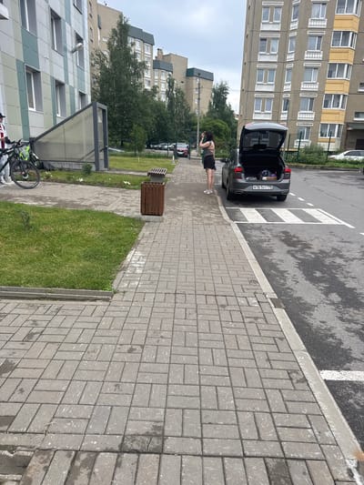 The image depicts an outdoor scene in the city of Bugry, Russia, featuring a paved sidewalk alongside a street. On the sidewalk, a woman is standing next to the open trunk of a dark-colored car, which contains several bags. A bicycle is parked on the grass adjacent to the sidewalk. In the background, several apartment buildings and trees are visible. The sky is overcast. The sidewalk is made of interlocking gray pavers, and there is a wooden trash can and a bench near the edge of the grass. The street has a pedestrian crossing marked with white stripes.