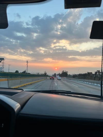 The photograph shows a view from inside a car driving on a highway at sunset. The sun appears as a bright red disc in the distance, obscured by haze. Cars are visible further down the road. The sky is filled with clouds, tinged with orange and blue hues from the setting sun. The interior of the car is visible on both sides of the frame, with parts of the dashboard and windshield present.