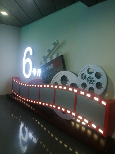 An indoor scene in Fengtai District, China, features a cinema-themed decorative display. Centrally positioned against a light blue-green wall is an illuminated white numeral "6" followed by the Chinese characters "号厅" (hào tīng), indicating "Hall 6." To the right and slightly behind, a brown-colored, stylized film clapperboard is visible, bearing the text "PROD: 光影看" (Guāng Yǐng Kàn) and fields for "SCENE," "TAKE," and "ROLL." Behind the clapperboard are two large, white, circular film reels. In the foreground, a curved, three-dimensional representation of a film strip with a red frame and grey sections exhibits numerous small, glowing white rectangular lights along its edges, simulating perforations. This entire display rests on a dark base. The dark, glossy floor below reflects the illuminated display elements. Overhead, a dark, paneled ceiling is partially visible, with one square light fixture reflecting on the floor. No individuals are present, and no actions are occurring.