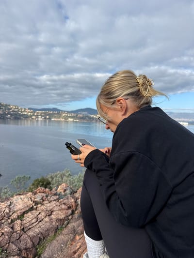 A blonde individual wearing glasses and a black long-sleeved shirt is seated on reddish-brown rock formations overlooking the Mediterranean Sea in Théoule-sur-Mer, France. The person's hair is pulled back with a gold-colored claw clip. They are engaged with a light-colored smartphone held in their right hand, while simultaneously holding a black vaping device in their left hand. A silver ring with a clear stone is visible on the ring finger of the left hand, and fingernails are painted a dark shade. The individual is also wearing black leggings, white ribbed socks, and light-colored athletic shoes.

The background features the calm, dark blue waters of a bay, with a distant coastline visible across the water. This coastline is dotted with numerous light-colored buildings and green vegetation, suggesting residential areas or small towns built into rolling hills. The sky above is largely covered with scattered, dense gray and white clouds, with patches of clear blue sky visible. Small green-grey shrubs are present on the rocky foreground to the left of the individual.