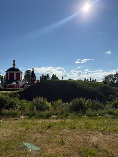 This is a photograph taken on a sunny day in Suzdal, Russia, likely at the Suzdal Kremlin or a similar historical site. The image depicts a vibrant red and white church with a golden dome, partially obscured by a large, grassy hill. Silhouettes of people are visible on top of the hill, standing and observing the surroundings. The sky is a clear blue with scattered white clouds, and the sun is bright and high, casting a strong glare. The foreground is dominated by dry, yellowish grass and lush green vegetation. The overall scene suggests a historical and scenic location with visitors enjoying the outdoor environment.