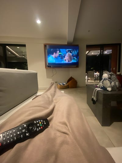 An indoor view shows a television mounted on a white wall. The TV is displaying a scene with two individuals in close proximity. Next to the TV is a decorative straw basket. A tan-colored soft chair or bed is in the foreground, with a black LG remote control lying on its surface. A plush doll is placed on a nearby surface in the background. The shot is taken from a slightly elevated position, providing a partial view of a window.